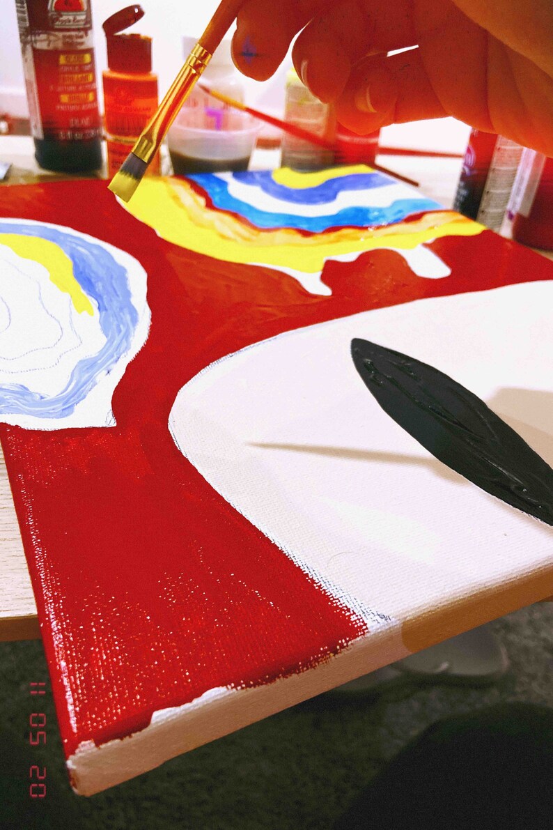 May include: A close-up of a canvas painting in progress. The painting features abstract shapes in red, white, blue, yellow, and black. A hand is holding a paintbrush and applying black paint to the canvas.