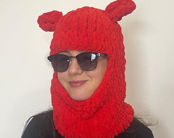 May include: A vibrant red balaclava-style hat with two small, rounded ear-like protrusions on top. The hat is made of a thick, textured material. The person is wearing black sunglasses and a black long-sleeve shirt.