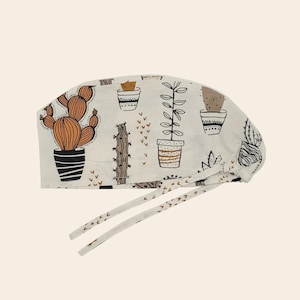 May include: A scrub cap in an off-white fabric featuring a repeating pattern of various potted cacti and succulents in shades of brown, black, and tan. The cap includes two long fabric ties at the back for securing the fit around the head.