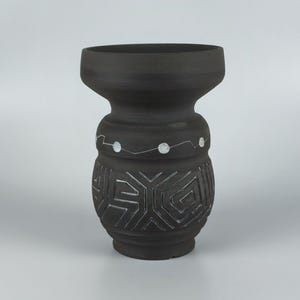 May include: A dark gray ceramic vase with a wide top and a bulbous midsection. The vase features white geometric patterns and dots. The design is reminiscent of tribal art. The vase is suitable for home decor.