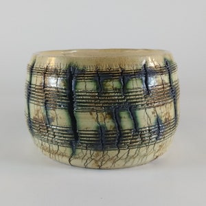 May include: A ceramic planter with a white glaze and a black and brown geometric pattern. The planter has a round shape and a wide opening.