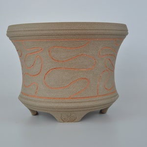 May include: A beige ceramic plant pot with a curved design and three small feet. The pot has orange, abstract, wavy lines around the body. The top and bottom rims have horizontal ridges. A decorative leaf design is on the base.