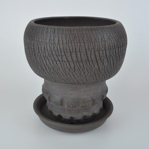 Peut inclure: Un pot en céramique gris foncé avec une surface texturée et une soucoupe assortie. Le pot a un haut large et arrondi et une base étroite.