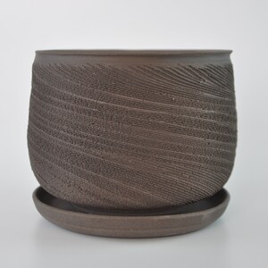 May include: A brown ceramic planter with a textured, swirling pattern. The planter has a rounded shape and sits on a matching saucer. The design gives the planter a rustic, handcrafted appearance, suitable for indoor or outdoor use.