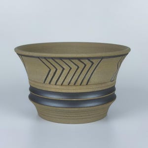 May include: A tan ceramic planter with a wide, flared rim. The planter features a dark brown chevron pattern around the top and two dark brown bands around the middle. The base is slightly rounded.