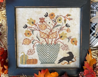 May include: A framed cross-stitch embroidery depicting a fall-themed floral arrangement in a green and white checkered pot. The arrangement features yellow, orange, and brown flowers, leaves, and stems. A black crow sits on a branch below the pot.