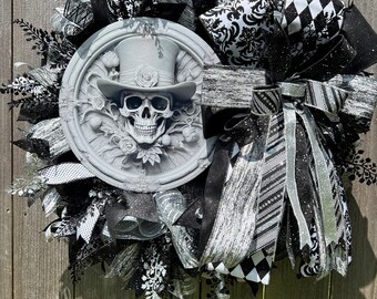 May include: Black and white Halloween wreath with a skull wearing a top hat in the center. The wreath is decorated with black and white ribbon, mesh, and other embellishments.