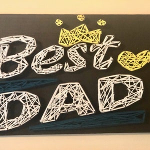 Puede incluir: Una pieza de arte de cuerda sobre un fondo negro con las palabras "Best Dad" en cuerda blanca. Una corona y un corazón amarillos están por encima de las palabras.