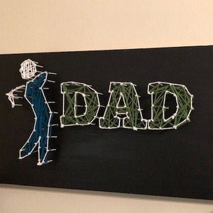 May include: A black wooden board with the word "DAD" spelled out in green and white string. A stick figure holding a golf club is also made out of string.