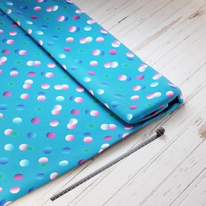 Könnte beinhalten: Gefalteter türkisfarbener Stoff mit einem sich wiederholenden Muster aus weißen, rosa und blauen Ovalen. Ein dunkelgraues Messwerkzeug liegt auf einer weißen Holzoberfläche neben dem Stoff.