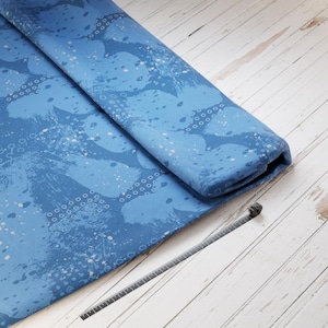 Könnte beinhalten: Gerollter blauer Stoff mit einem Aquarelldesign in Blau- und Weißtönen. Ein graues Maßband liegt auf einer weißen Holzoberfläche.