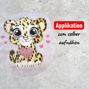 Könnte beinhalten: Ein gesticktes Applikationsmotiv mit einem Cartoon-Leopardenbaby mit großen Augen und einem rosa Glitzerherz. Der Leopard hat ein geflecktes Fell und ist von kleinen rosa Herzen umgeben. Text in einem roten Feld: "Applikation zum selber aufnähen."