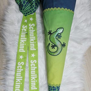 Könnte beinhalten: Grüner und blauer Schultüte mit einer grünen Eidechsen-Applikation und dem Text "Schulkind" wiederholt auf zwei grünen Bändern.