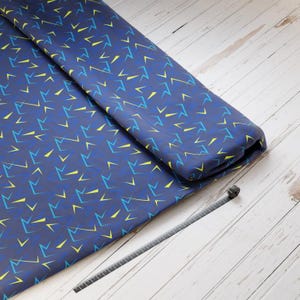 Könnte beinhalten: Ein blauer Stoff mit einem sich wiederholenden Muster aus gelben und blauen geometrischen Formen. Der Stoff ist gefaltet und liegt auf einer weißen Holzoberfläche.