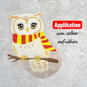 Könnte beinhalten: Ein Eulen-Applikation mit weißem Körper, goldenen Akzenten und einem rot-gelb gestreiften Schal. Die Eule sitzt auf einem braunen Ast. Text in roter Box: "Applikation zum selber aufnähen".