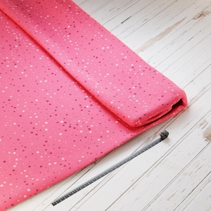 Könnte beinhalten: Eine gefaltete Rolle aus rosa Stoff mit einem gesprenkelten Muster aus weißen und dunkelrosa Punkten. Ein graues Maßband liegt neben dem Stoff auf einer weißen Holzoberfläche.