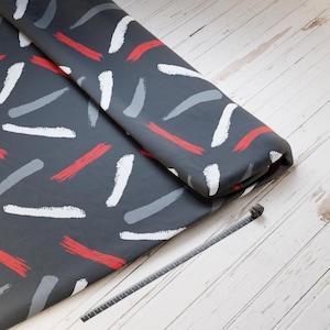 Könnte beinhalten: Dunkelgrauer Stoff mit einem Muster aus weißen, roten und grauen Pinselstrichen. Der Stoff ist gerollt, und ein schwarzer Kabelbinder liegt auf einer hellen Holzoberfläche.