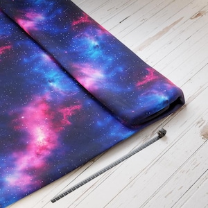 Könnte beinhalten: Nahaufnahme eines dunkelblauen Stoffes mit einem Galaxie-Druck, der rosa, blaue und weiße Nebel und Sterne zeigt. Der Stoff ist aufgerollt und ein Maßband liegt auf einer weißen Holzoberfläche.