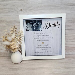 May include: A white framed print with the word "Daddy" in cursive. The print features a sonogram image and a poem about a baby's love. A small white vase with dried flowers is to the left of the frame.