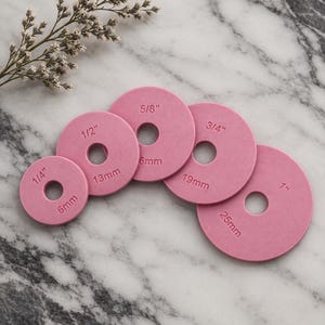 Op de afbeelding: Een set van vijf cirkelvormige roze schijven met gaten in het midden, elk voorzien van metrische maten van 6mm, 13mm, 6mm, 19mm en 25mm, naast imperiale maten van 1/4 inch, 1/2 inch, 5/8 inch, 3/4 inch en 1 inch op het oppervlak.
