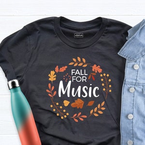 May include: A dark gray t-shirt with the text "FALL FOR Music" surrounded by a wreath of autumn leaves. A blue and orange water bottle and a light blue denim jacket are also in the image.