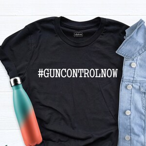 May include: Black t-shirt with the text "#GUNCONTROLNOW" in white lettering. The t-shirt is displayed on a white surface with a denim jacket and a water bottle.