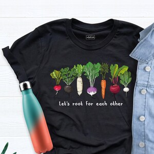 May include: Black t-shirt with a graphic of a row of colorful vegetables, including carrots, radishes, turnips, and beets. The text "Let's root for each other" is printed below the vegetables.