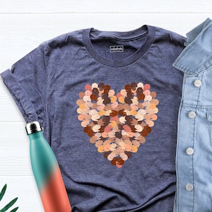 May include: A navy blue t-shirt with a heart-shaped design made up of many hands of different skin tones. The t-shirt is laid flat on a white surface with a denim jacket and a water bottle.