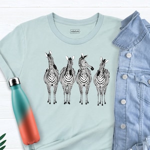 May include: A light blue t-shirt with a graphic of four zebras standing in a row. The zebras are black and white with a striped pattern. The t-shirt is on a white surface with a denim jacket and a water bottle.