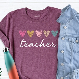May include: A maroon t-shirt with the word "teacher" in white script, above five colorful heart designs. A light blue denim jacket is draped over the shirt. A teal and orange water bottle is on the left.