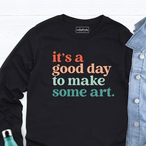 May include: Black long-sleeve t-shirt with the text "it's a good day to make some art." in a colorful gradient design.