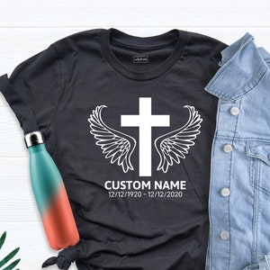 May include: Black t-shirt with a white cross and angel wings design. The text "CUSTOM NAME 12/12/1920 - 12/12/2020" is printed below the design.