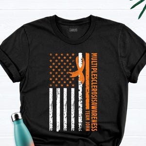 May include: Black t-shirt with an orange ribbon and an American flag design. The text "Multiple Sclerosis Awareness Team John" is printed on the shirt.