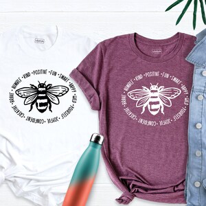 May include: Two t-shirts with a black and white bee graphic and a circle of positive affirmations around it. The text reads: "Brave, Humble, Kind, Positive, Fun, Smart, Happy, Wild, Yourself, Joyful, Confident, Creative".