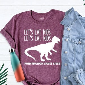 May include: Burgundy t-shirt with a white dinosaur graphic and the text "Let's eat kids. Let's eat, kids. Punctuation saves lives."