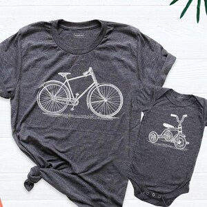 May include: A matching set of grey t-shirts featuring a white bicycle and a white tricycle. The adult t-shirt has a crew neck and the baby t-shirt has a snap closure.