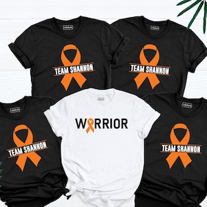 May include: Five t-shirts, four black and one white, are displayed. The black shirts feature an orange ribbon and the text "TEAM SHANNON". The white shirt has the word "WARRIOR" with an orange ribbon. The shirts are short-sleeved.