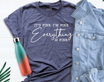 Könnte beinhalten: Ein marineblaues T-Shirt mit dem Text "It's fine. I'm fine. Everything is fine." in weißen Buchstaben. Das Shirt liegt flach auf einer weißen Oberfläche mit einer Jeansjacke und einer Wasserflasche.