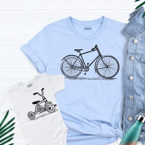 May include: A light blue t-shirt with a black and white illustration of a bicycle. A white baby onesie with a black and white illustration of a tricycle. A denim jacket and a water bottle are also in the image.