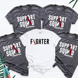 May include: Four gray t-shirts with white text and a red ribbon design. The shirts say "Support Squad #TeamEmma" and "Fighter".
