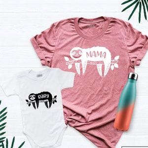 May include: A white baby onesie with a black sloth graphic and the word "baby". A dusty rose t-shirt with a white sloth graphic and the word "MAMA". A teal and orange ombre water bottle.