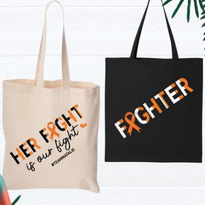 May include: Two tote bags with orange ribbon designs. One bag is white with the text "HER FIGHT is our fight #TEAMNATALIE". The other bag is black with the text "FIGHTER".