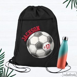 May include: Black drawstring backpack with a soccer ball graphic and the name "Jackson" in red. The soccer ball has the number 17 in red. A teal and orange gradient water bottle is next to the bag.