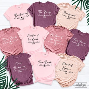 May include: Nine different t-shirts with various wedding party titles and names printed on them. The shirts are in pink, peach, and burgundy colors.