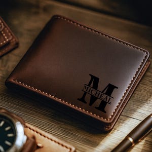 May include: A brown leather wallet with the name "Micheal" and the letter "M" engraved on the front. The wallet is surrounded by a watch, pen, and another wallet on a wooden surface.