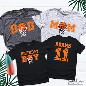 May include: Four short-sleeve t-shirts with basketball-themed graphics. One dark gray shirt says DAD, one light gray says MOM, one black says BIRTHDAY BOY, and one black says ADAMS 11. All feature orange basketballs and white nets.
