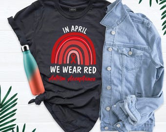 May include: A black t-shirt featuring a red rainbow graphic with a red ribbon and the text IN APRIL WE WEAR RED Autism Acceptance, paired with a light blue denim jacket and a teal and orange gradient metal water bottle.