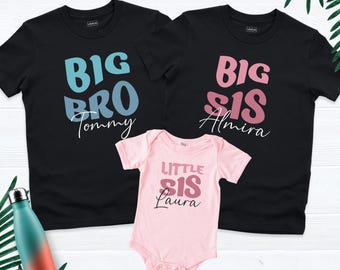 May include: Two black t-shirts and one light pink baby bodysuit. The first black shirt features blue text reading BIG BRO Tommy. The second black shirt has pink text reading BIG SIS Almira. The pink bodysuit features dark pink text reading LITTLE SIS Laura.