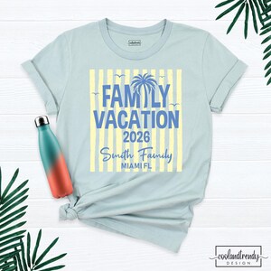 May include: A light blue t-shirt featuring a graphic with yellow vertical stripes, a blue palm tree, and the text "FAMILY VACATION 2026 Smith Family MIAMI FL". Beside the shirt is a teal and orange gradient metal water bottle.