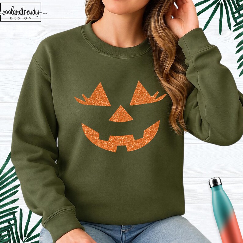 May include: Olive green sweatshirt featuring a glittery orange jack-o'-lantern face. The design includes triangular eyes, a triangular nose, and a smiling mouth. The sweatshirt has a crew neck and long sleeves. The text "coolandtrendy DESIGN" is in the upper left corner.